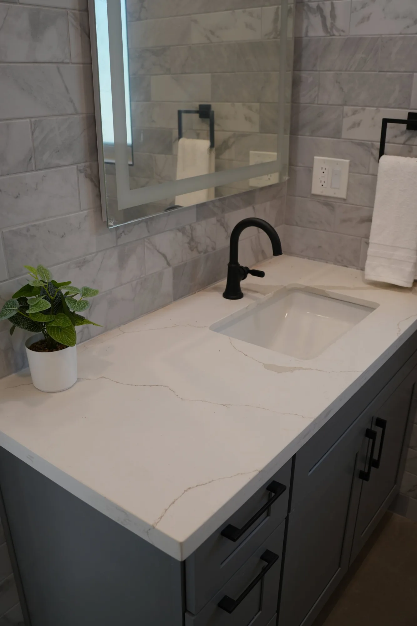 Gray vanity with a white veined countertop, black faucet and lighted mirror